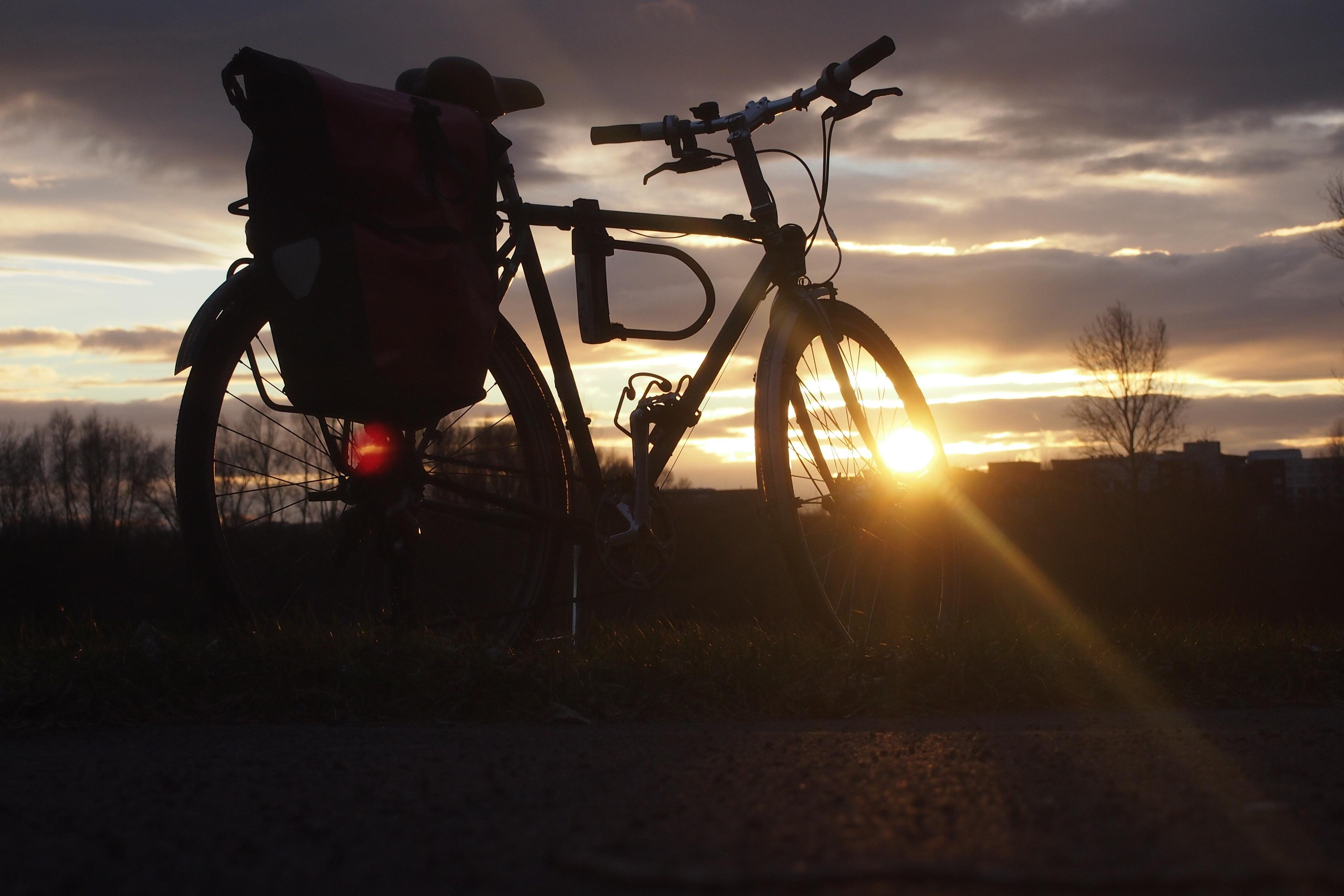 Foto im Querformat. Die Kamera befindet sich knapp über dem Boden. Im Vordergrund steht ein Fahrrad mit einer roten Fahrradtasche. Durch das Vorderrad scheint die untergehende Sonne, Sonnenstrahlen reichen bis in die rechte untere Ecke. Im Hintergrund befinden sich blätterlose Bäume und Häusersiloutten. Der Himmel ist mit dunklen Wolken bedeckt, ihre Kanten sind durch das Sonnenlicht hervorgehoben.